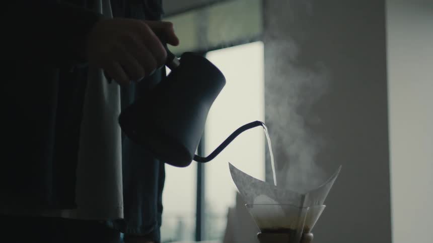 A person is pouring hot water from a gooseneck kettle into a coffee maker with steam rising from the filter