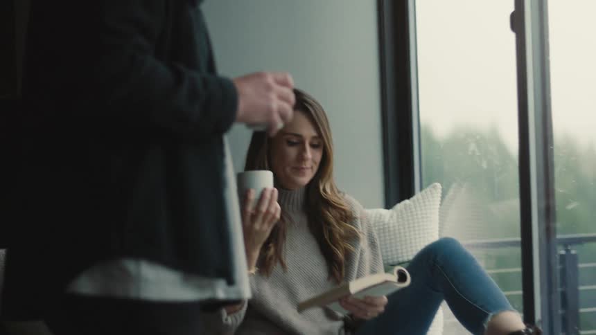 A man brings a cup of coffee to a woman sitting on a couch by a large window holding a book