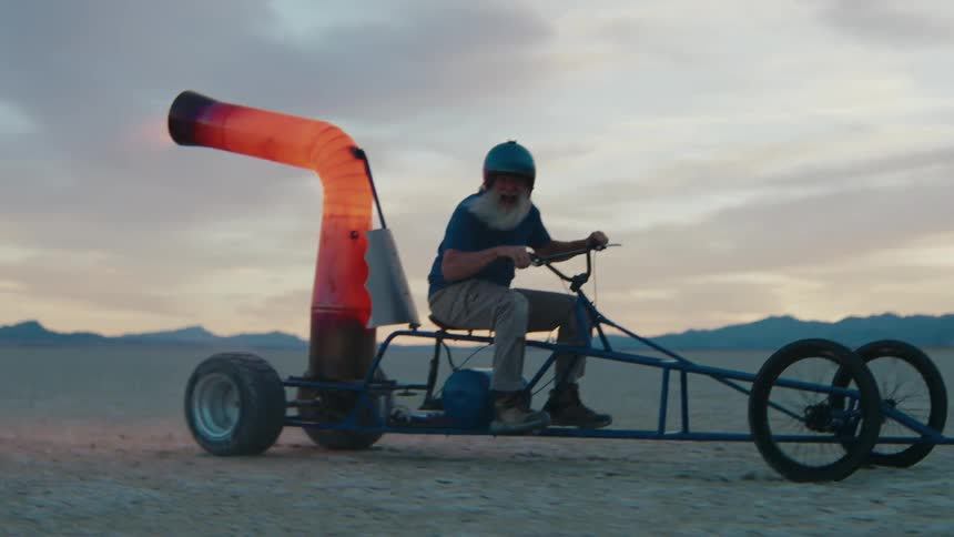 Older man rides a cart powered by thrust across a desert under a red tinted sky