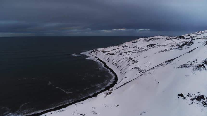 A snowcovered hillside slopes down to a dark rugged coastline bordering a turbulent sea under a cloudy sky