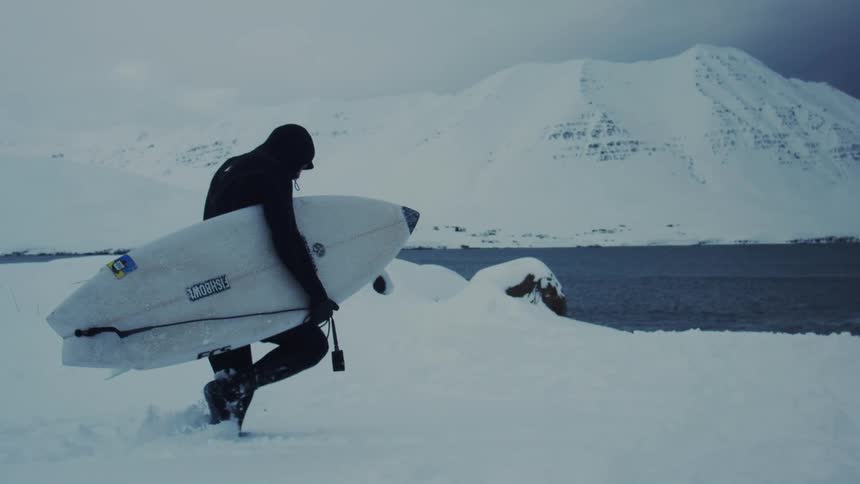 Person in a swimsuit holding a surfboard walking towards the ocean in a snowy landscape