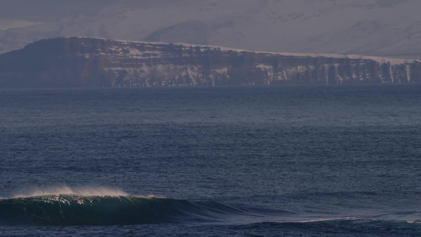 A small wave breaks in the foreground of a vast rippling ocean with a large flattopped mountain looming in the hazy distance