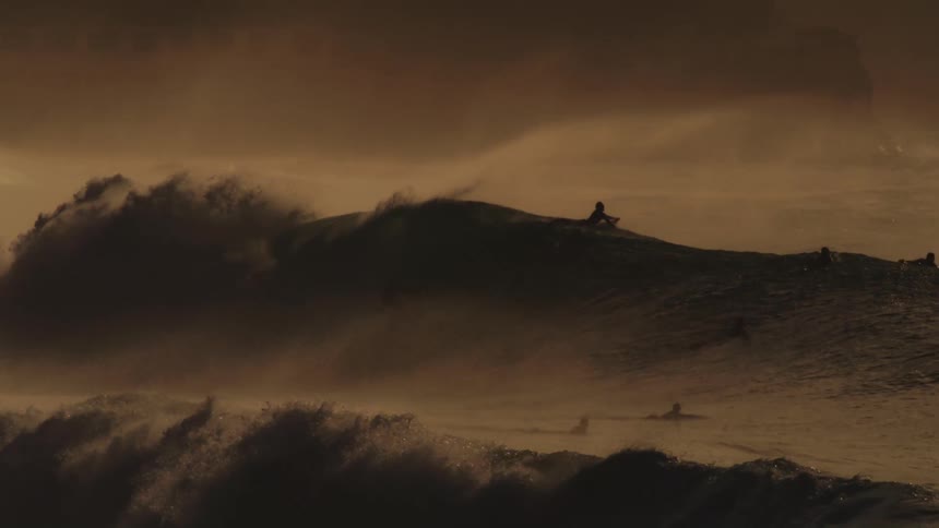 A massive wave crashes in a swirl of foam and spray illuminated by a warm golden light with surfers dwarfed by the size of the wave
