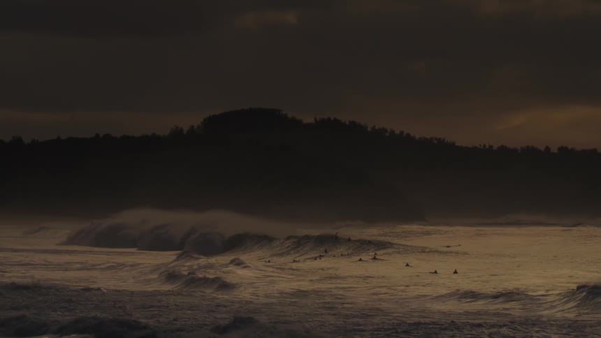 Waves roll onto a sandy shore under a dark dramatic sky with a silhouetted hill in the distance