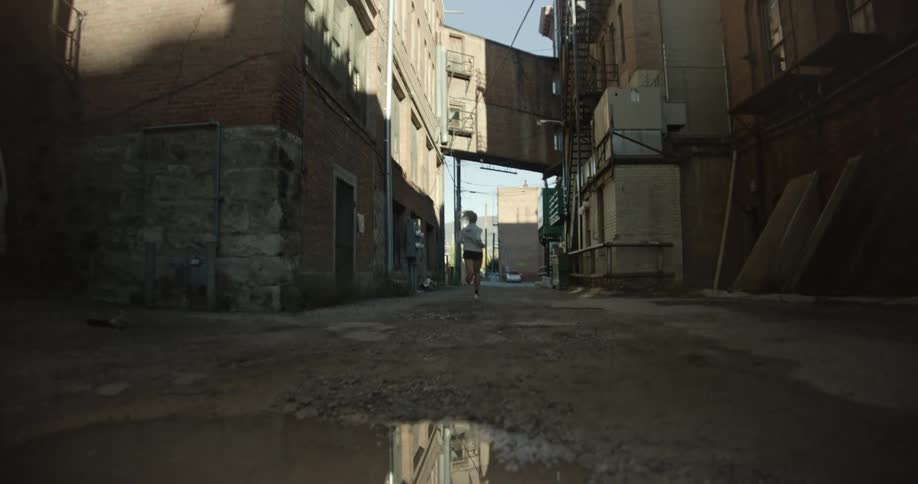 A woman is running down a narrow dimly lit alleyway with a large puddle in the foreground reflecting the surrounding buildings and the runner