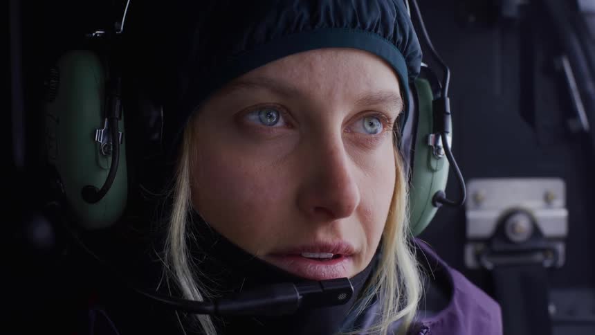 A blonde woman with blue eyes and a focused expression wears a dark cap and headphones with a microphone