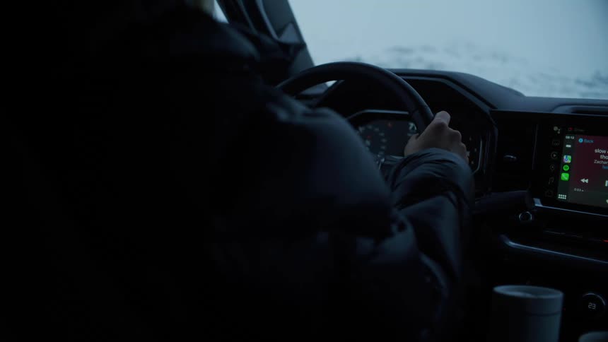 A person is driving a vehicle in a snowy environment with the dashboard displaying a music playlist on the screen