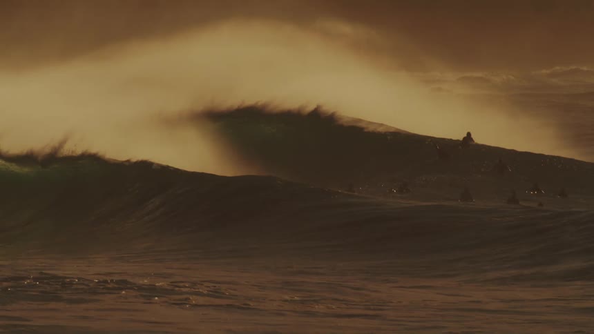 A massive wave crashes in a swirl of foam and spray illuminated by a warm golden light against a hazy sky with surfers dwarfed by the size of the wave