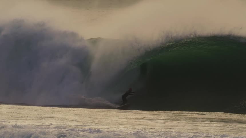 A surfer rides a large dark wave that curls and breaks in the ocean leaving a trail of haze