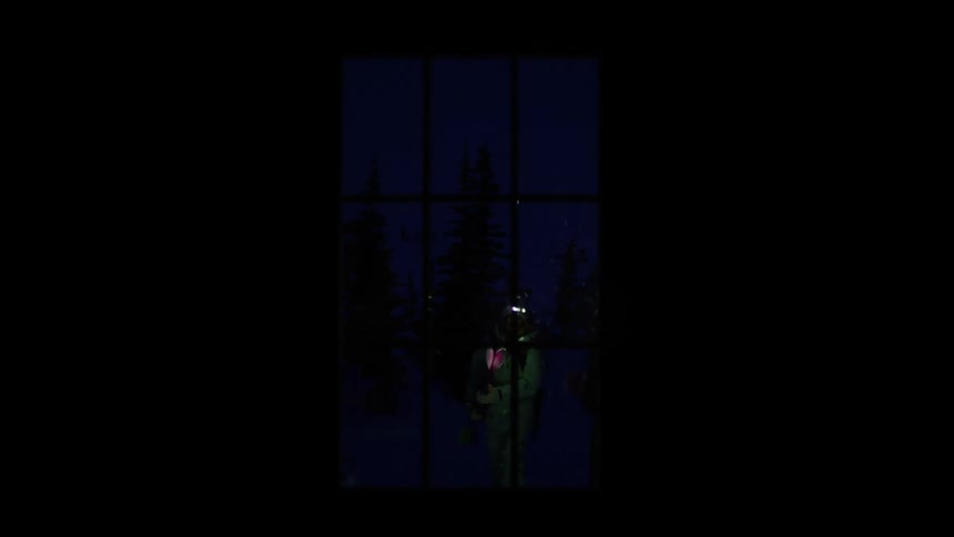 A nighttime view through a window showing two people with headlamps and winter clothes approaching the scene
