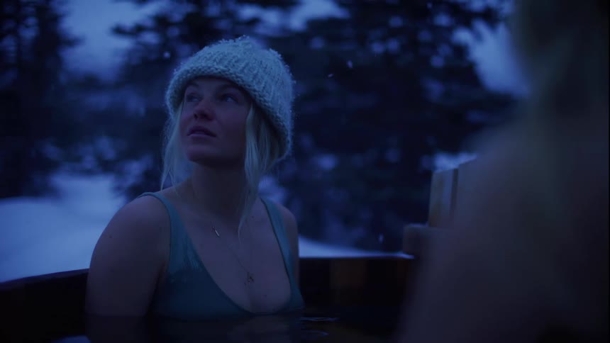 Woman in beanie and bathing suit stands inside outdoor wooden bathtub surrounded by snow and trees at night
