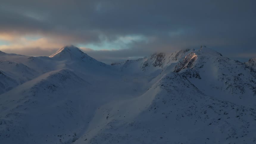 Snowcovered mountain peaks rise into a dramatic sky with patches of sunlight breaking through storm clouds