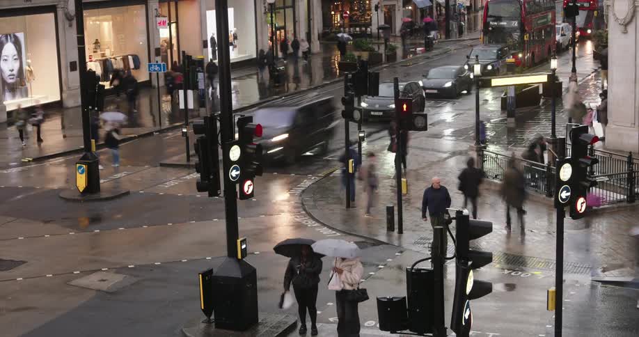 A busy city street intersection with red doubledecker bus pedestrians holding umbrellas and wet pavement indicating recent rain