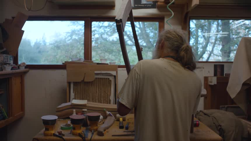In a woodshop with two windows a man with long hair works on a wooden chair at a workbench littered with tools