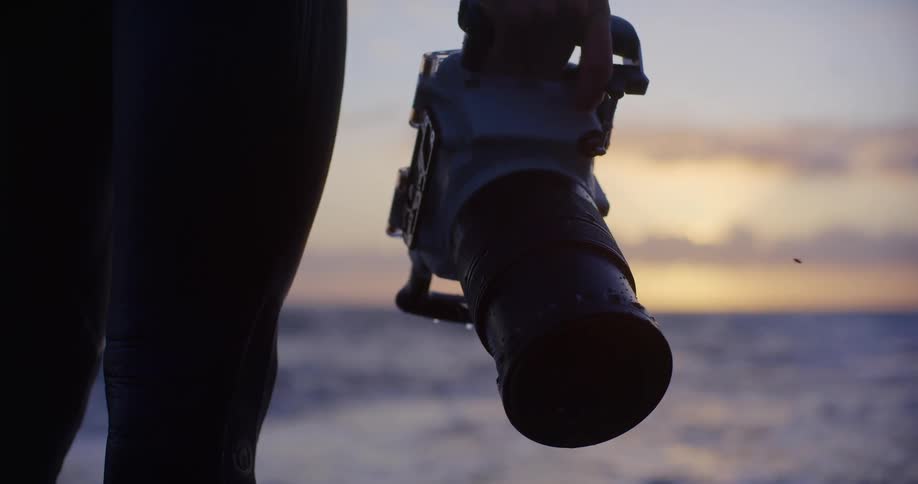 Male photographer wearing a wet suit stands on the shore holding a camera