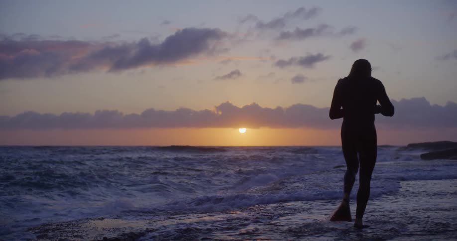 Man wearing a wet suit and flippers walks through the surf on the coast near sunset
