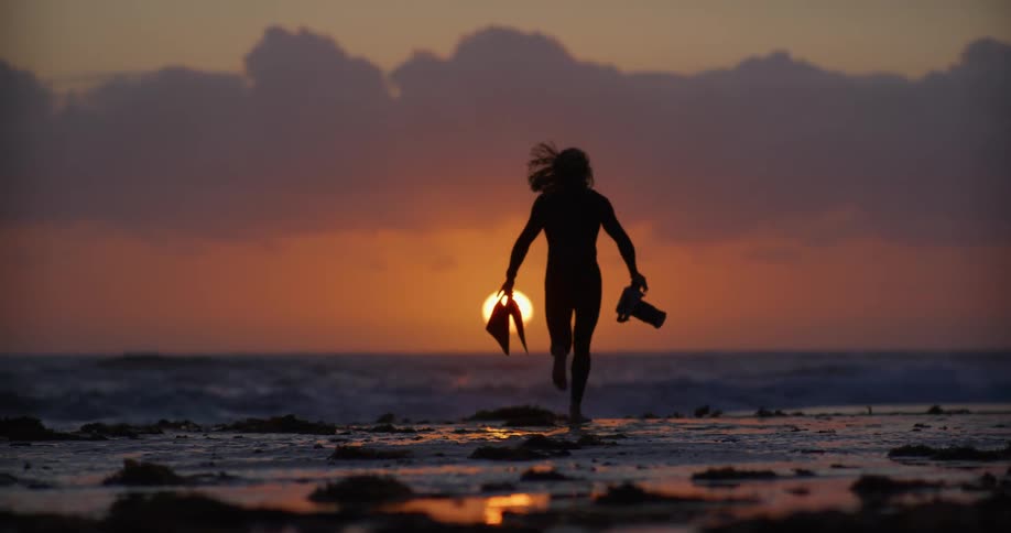 Man wearing a wet suit carries flippers and a camera while running towards the surf and the setting sun