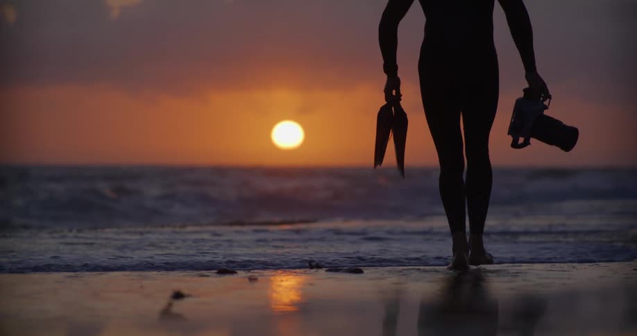 Silhouette photographer holding camera and fins running in ocean at sunrise
