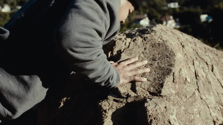 A person wearing a gray sweatshirt and a black beanie steadies themselves with a hand on a rock overlooking houses on a hill