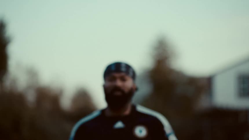 A bearded man in a chelsea soccer jersey and bandana runs towards the camera talks and then runs out of frame outdoors