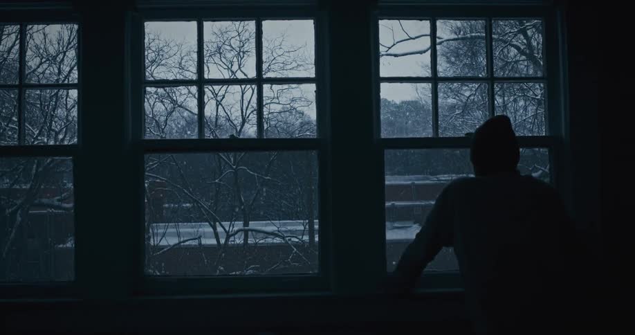 A hooded silhouette of a person stands by a window watching the snowfall outside
