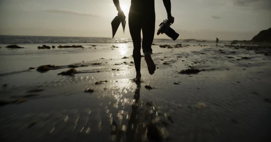 Silhouette of a person carrying a professional camera and flippers running towards the sea