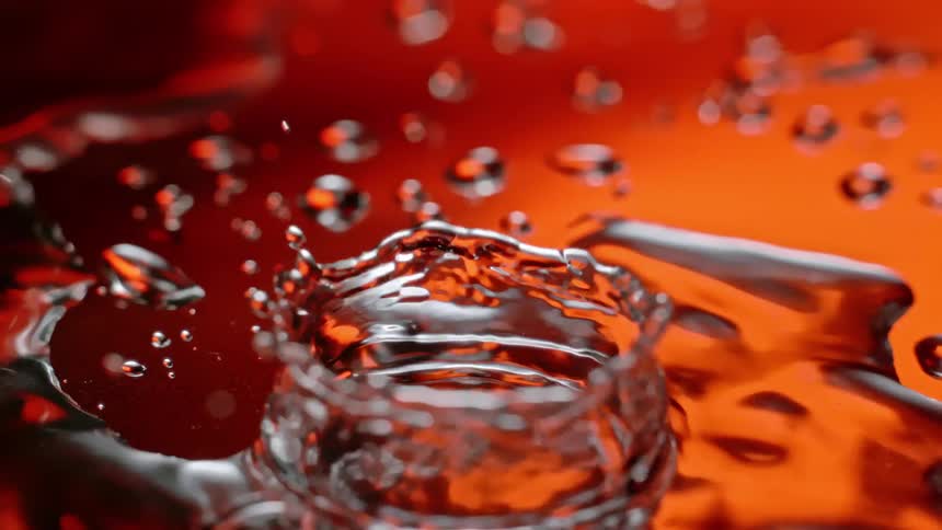 A drop of liquid falls into a pool of liquid creating a splash and ripples against an orange background