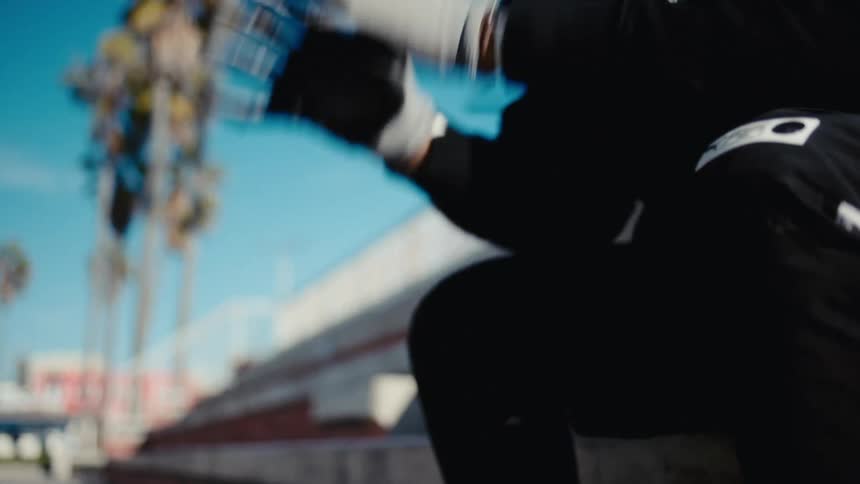 A person in a black tracksuit putting on sports gloves dribbles a soccer ball with palm trees and a blue sky blurred in the background