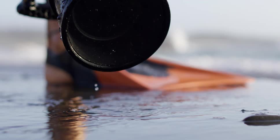 A person with fins lifts a dripping camera lens out of shallow water on a beach causing ripples
