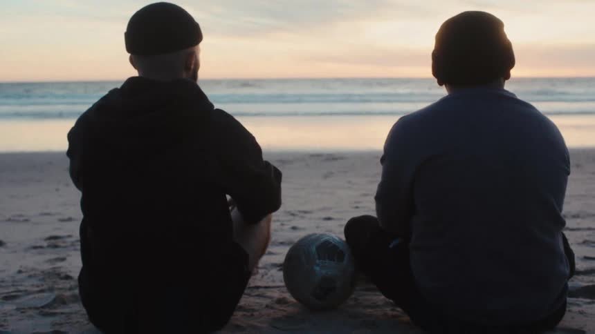 Two friends sitting on the sand watching a dog run along the shoreline at sunset