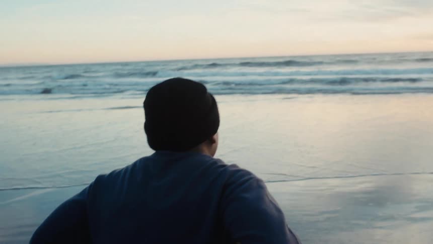 Man bends over to rest while on a beach of wet reflective sand