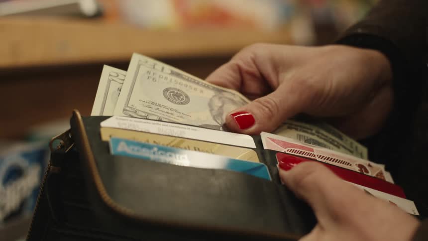 A close up shows a persons hands with red nail polish pulling a twenty dollar bill from a black wallet filled with cash and credit cards
