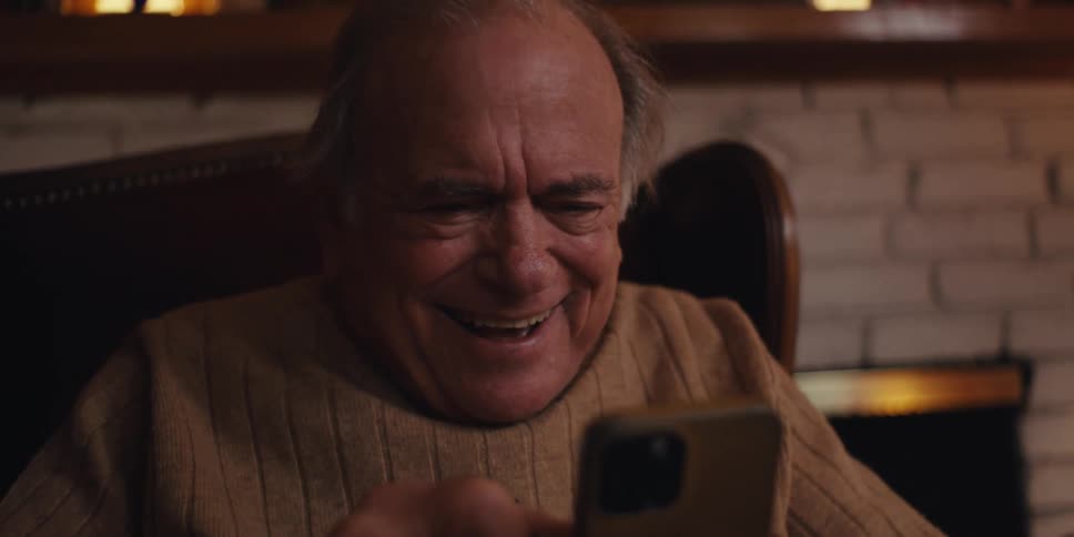 An older man smiles broadly while looking at his phone indoors