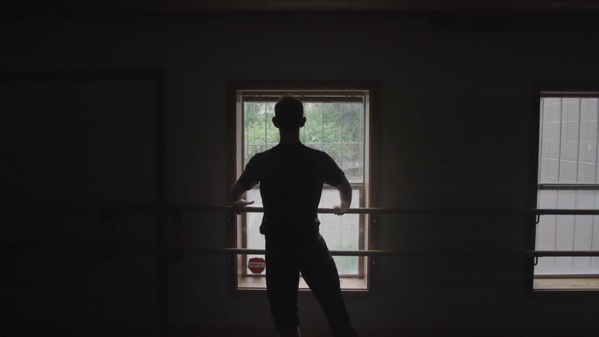A man is silhouetted against a window reclined on a ballet barre looking out of the window from a dimly lit studio