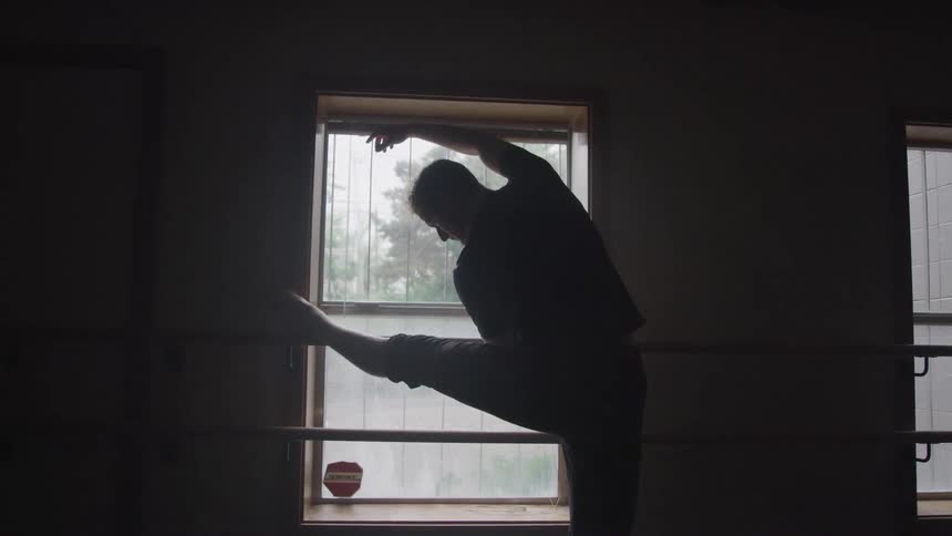 A man is performing a ballet stretch with one leg extended on a barre in front of a window creating a silhouette against the light coming through the window