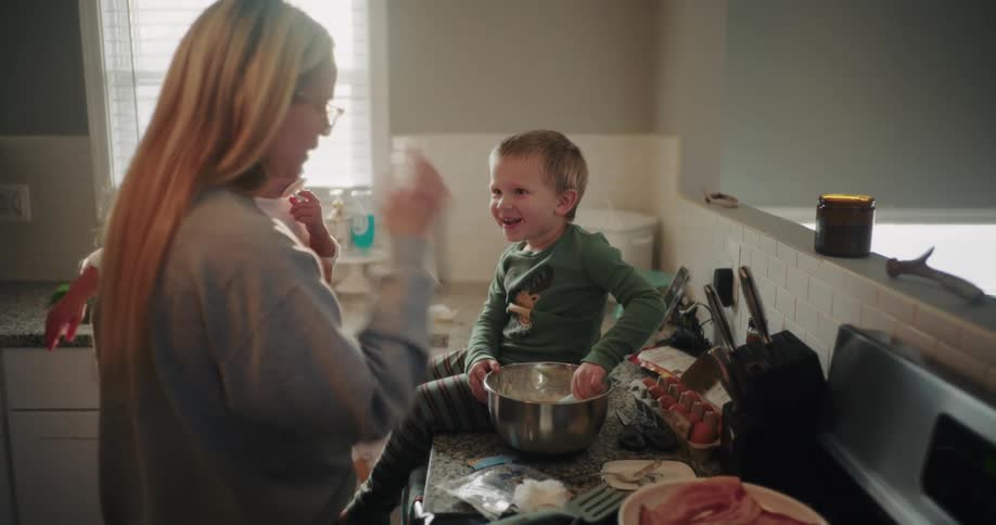 Mother holds a baby while her toddler son sits on the counter tasting cookie dough