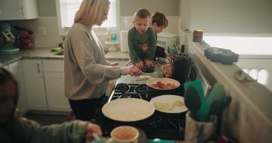 In a brightly lit kitchen a woman cooks at the stove while two young boys sit on the counter and a young girl pours batter into a bowl