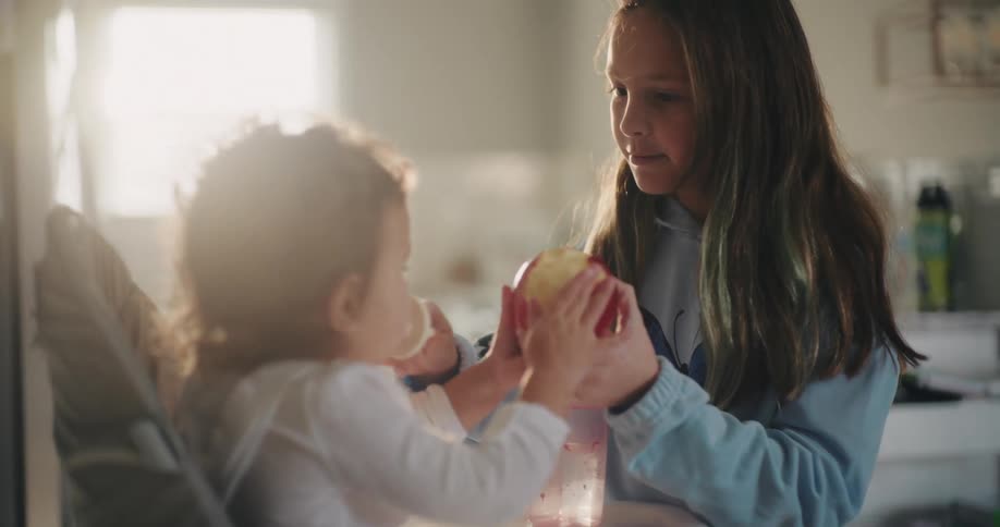 A baby is sitting in a high chair while her sister wearing a light blue top talks to her and hands over apples and a drink