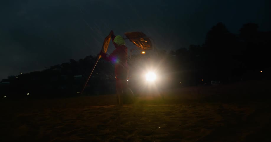 A lifeguard plants a sign on a sandy beach illuminated by the headlights of a small vehicle at sunset