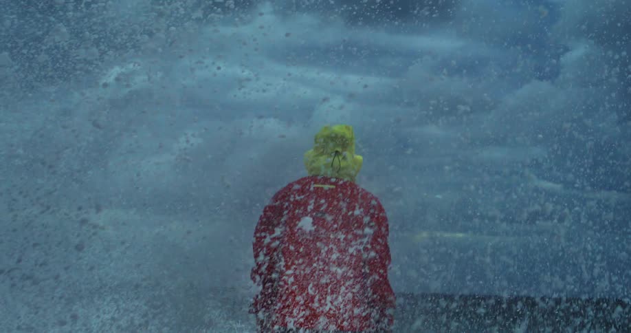 A person in a red outfit and yellow hat stands on a rocky shore facing a massive wave crashing against the rocks under a stormy sky