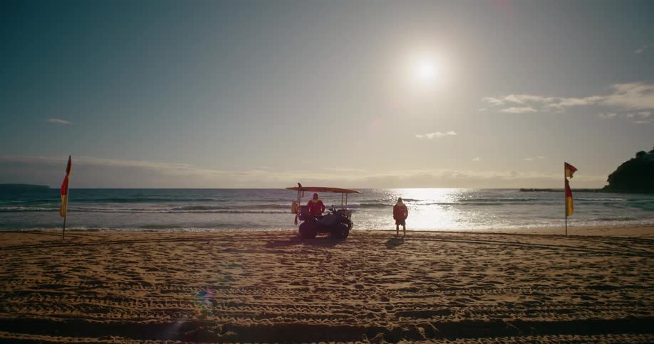 Lifeguards stand on the beach with an atv with surfboard on top of a cargo rack flags wave as ocean waves reach the shore in sunny daylight