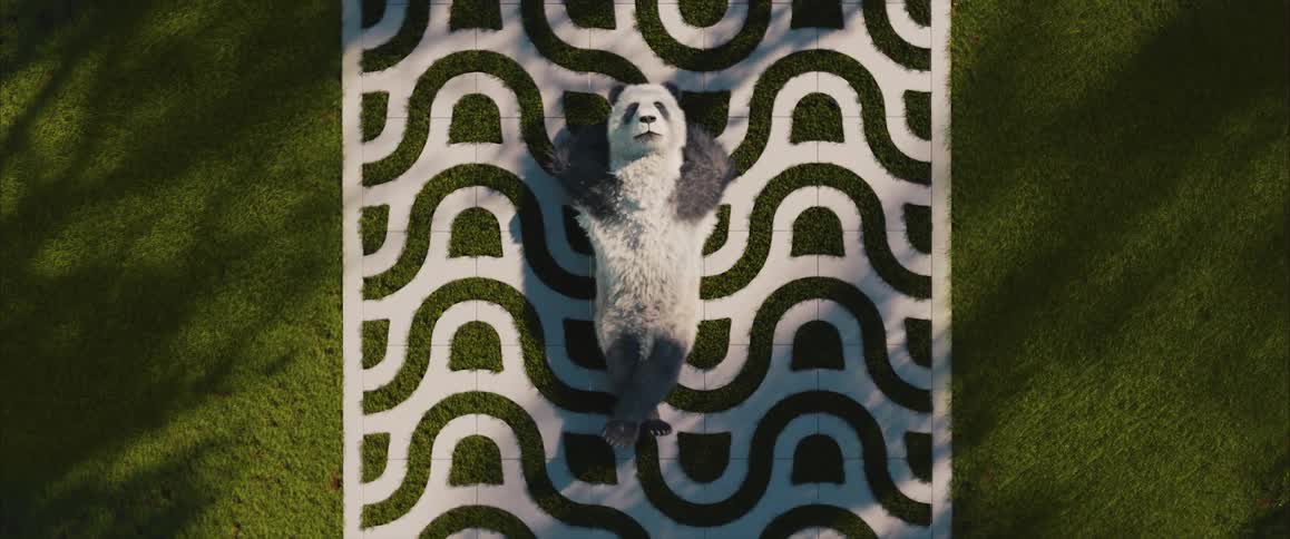 A panda lies on its back on a geometrically patterned rug surrounded by green grass viewed from directly above