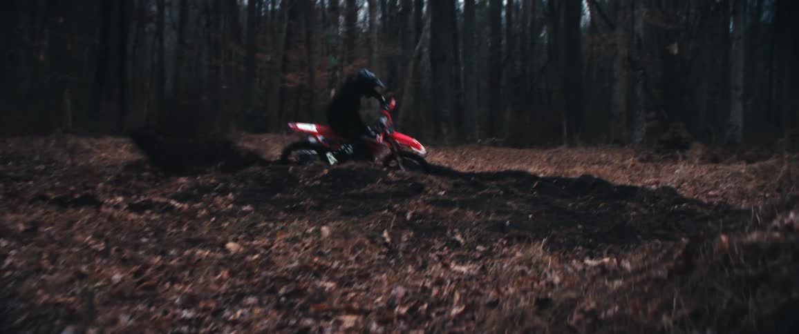 A person wearing a helmet is riding a red dirt bike through a dense forest spinning around and falling