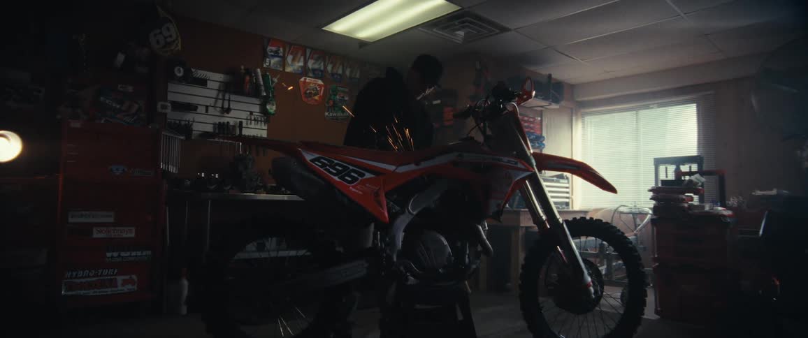 A person is using a power tool to grind or cut metal on a motorcycle causing bright sparks to fly in a workshop filled with various tools and equipment