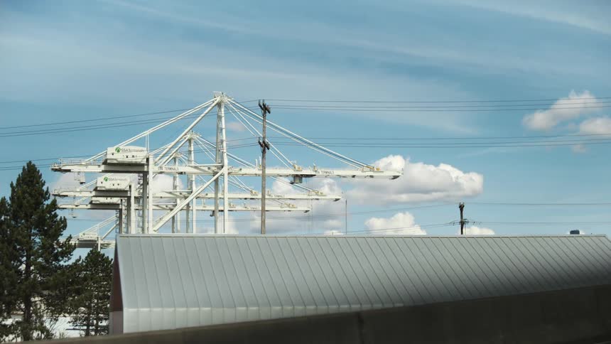 A white gantry crane structure stands against a blue sky with scattered clouds partially obscured by trees and a building with a gray metal roof