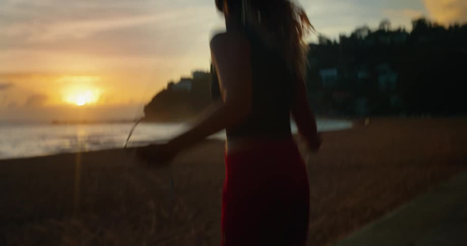 A woman is jumping rope on a beach at sunset with the sun low on the horizon and a silhouette of a hilly area with houses in the background