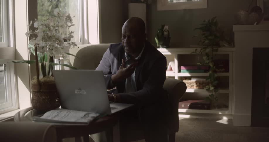A man is sitting in an armchair using a laptop in a room and he is talking to the screen