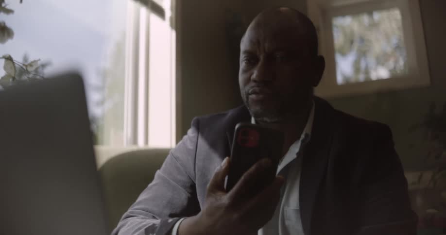 Inside a living room a black man in a blazer holds an iphone to his ear engaged in a call while working on a laptop