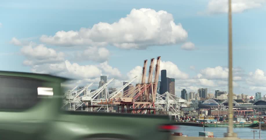 A busy port with large white cranes lifting and moving stacked shipping containers seen from a highway