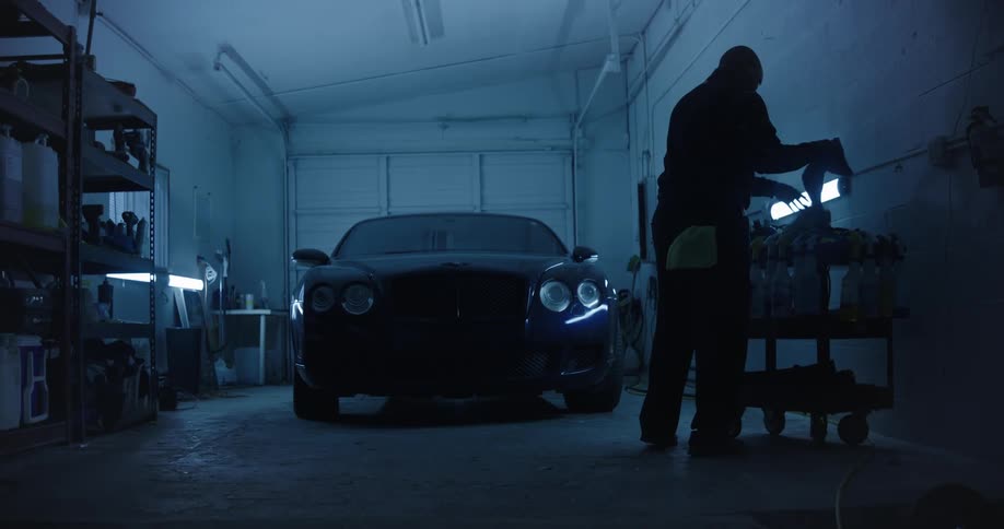 A person is working on a car in a dimly lit garage with various tools and equipment visible around the space